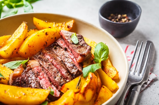 Gegrilde biefstuk met aardappelen en basilicum in een witte plaat op witte achtergrond.