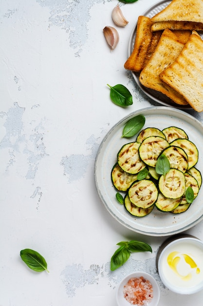 Gegrilde courgettesalade met basilicumblaadjes, yoghurtsaus en gebakken brood in een eenvoudige keramische plaat op een witte betonnen ondergrond