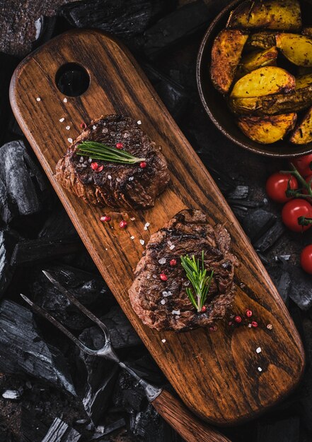 Gegrilde filetlapjes vlees op houten scherpe raad. sappige dikke sappige porties van gegrilde haasbiefstuk geserveerd met tomaten en gebakken aardappelen op een oude houten plank. bovenaanzicht.