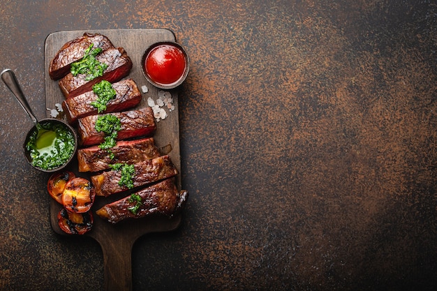 Gegrilde/gebakken en gesneden gemarmerde vlees steak met vork, tomaten, verschillende sauzen op houten snijplank, bovenaanzicht, close-up met ruimte voor tekst, rustieke stenen achtergrond. Rundvlees steak concept