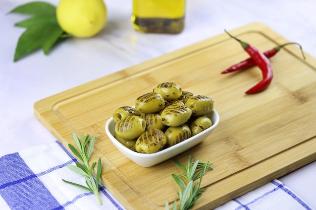 Gegrilde groene olijven met rozemarijn, chilipeper, citroen en olie in een bord op snijplank op tafel