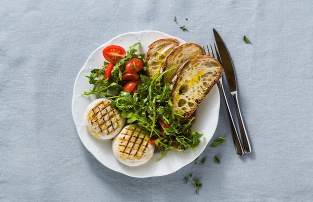 Gegrilde Italiaanse Tomino kaas geserveerd op een tafel met rucola salade en verse zelfgemaakte ciabatta brood en tomaat op een blauw linnen feestelijk tafelkleed. zomer menu