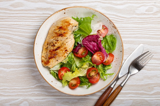 Gegrilde kipfilet met groentensalade van verse tomaten en saladebladeren op witte plaat op houten ondergrond van bovenaf, gezond diner of dieetmaaltijd
