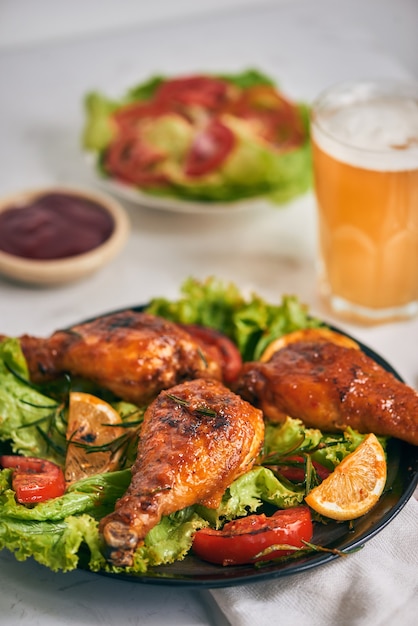 Foto gegrilde kippenpoten geroosterd op de grill op donkere plaat met tomatensaus in een kom en slabladeren, glazen mok bier