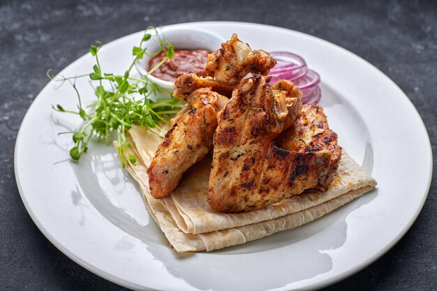 Gegrilde kippenvleugels met barbecuesaus, pitabroodje, microgreen en uienringen, op een witte plaat, tegen een donkere tafel