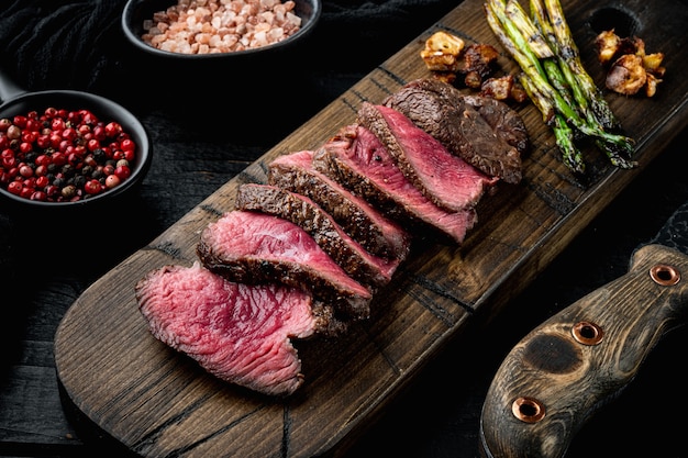 Gegrilde ossenhaas steaks set, met ui en asperges, op houten serveerplank, met vleesmes en vork, op zwarte houten tafel achtergrond