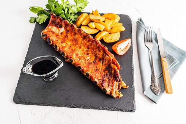 Gegrilde varkensribbetjes op een leisteen donker bord met plakjes aardappelen tomatensaus gastronomische lunch heerlijk eten witte achtergrond