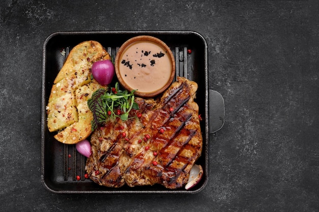 Gegrilde varkenssteak met aardappelen, kruiden en saus op een donkere achtergrond in een koekenpan