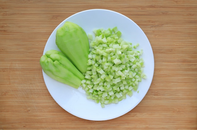 Gehakte en gesneden Chayote op witte plaat tegen houten raadsachtergrond.