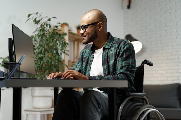 Gehandicapte jonge Afro-Amerikaanse man in rolstoel die computer gebruikt terwijl hij aan zijn werktafel zit