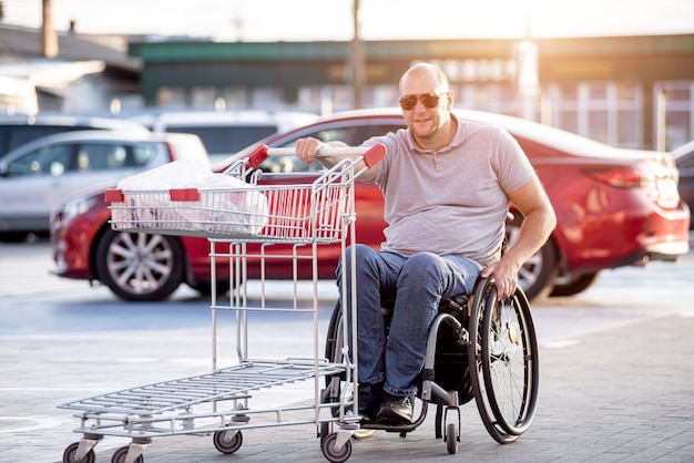 Gehandicapte man in rolstoel die kar voor zich uit duwt bij supermarktparkeren