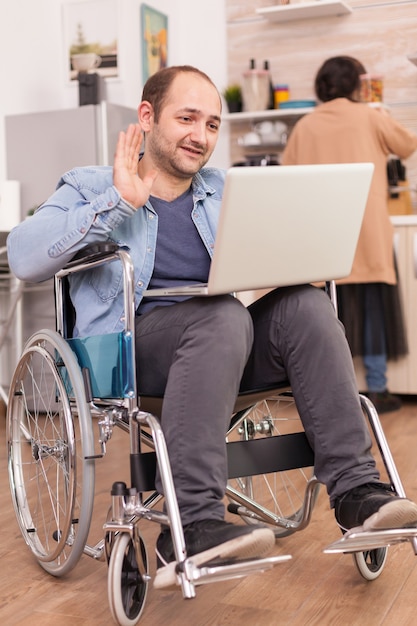 Gehandicapte ondernemer in rolstoel die tijdens een videogesprek op laptop zwaait terwijl vrouw lunch kookt. Gehandicapte verlamde gehandicapte man met loopbeperking integreren na een ongeval.