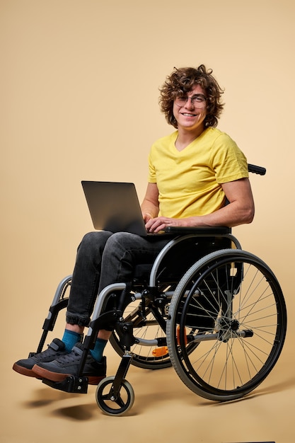 Gehandicapte persoon in vrijetijdskleding zittend met laptop op rolstoel, geïsoleerd op beige studio achtergrond. knappe gekrulde man geniet van online werken, bericht typen op toetsenbord