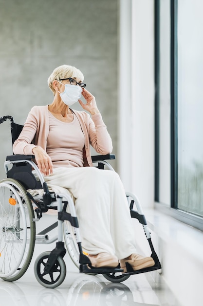 Gehandicapte senior vrouw met beschermend masker zittend in een rolstoel en peinzend uit het raam kijkend tijdens de pandemie van het coronavirus.