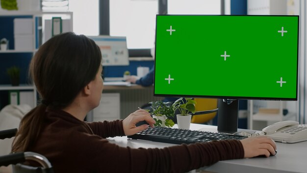 Gehandicapte vrouwelijke ondernemer die op een desktopcomputer werkt met groene Chromakey, projectinformatie typt terwijl hij in een rolstoel zit