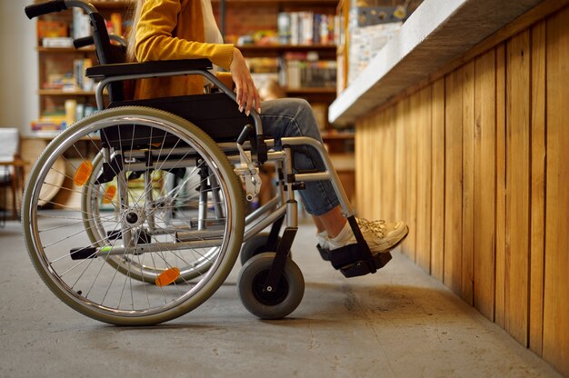 Gehandicapte vrouwelijke student in rolstoel aan de balie, handicap, boekenplank en universiteitsbibliotheek of café interieur op de achtergrond. Gehandicapte vrouw op de universiteit, verlamde mensen krijgen kennis