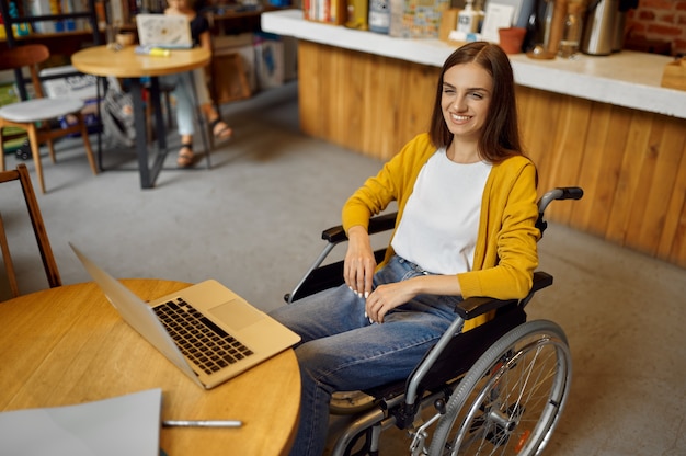 Gehandicapte vrouwelijke student in rolstoel met behulp van laptop, handicap, boekenplank en universiteitsbibliotheek interieur op de achtergrond. Gehandicapte jonge vrouw studeert op de universiteit, verlamde mensen