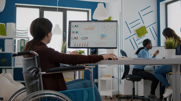 Gehandicapte zakenvrouw die financiële statistieken analyseert die in een rolstoel zitten en grafieken van de computer in het startbureau controleren. Gehandicapte werknemer die met een team werkt dat moderne technologie gebruikt