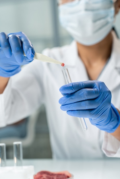 Gehandschoende handen van hedendaagse onderzoeker in witte jas en masker die een klein stukje rauw plantaardig vlees in de fles stopt tijdens laboratoriumexperiment