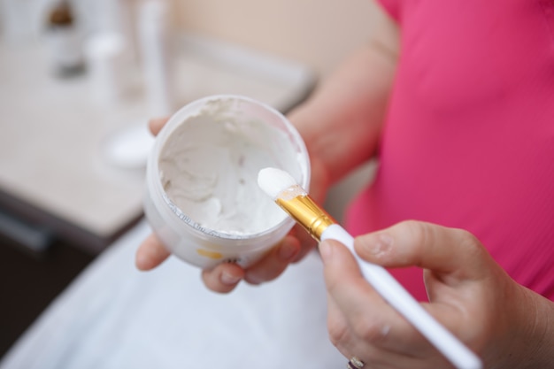 Gehandschoende schoonheidsspecialiste die gezichtscrème of masker met borstel neemt voordat het op het gezicht van de klant wordt aangebracht