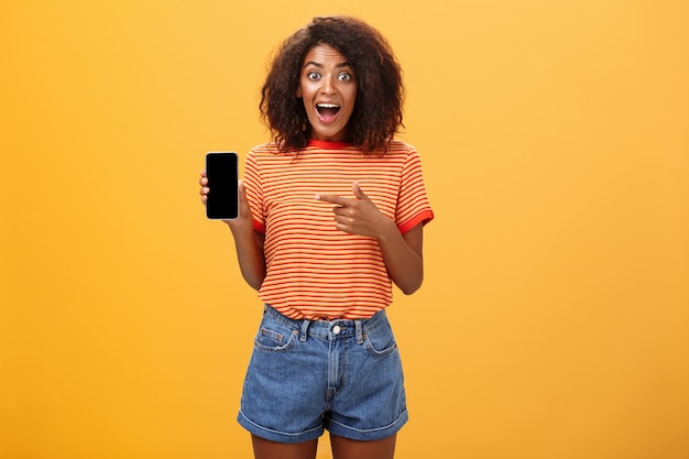 Geïmponeerde Afro-Amerikaanse vrouw met krullend haar wijzend op mobiel over oranje muur