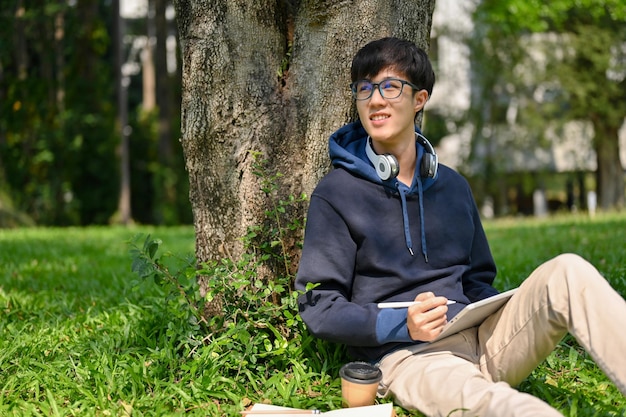 Geïnspireerd en gelukkig jonge Aziatische mannelijke student zittend onder de boom in het park