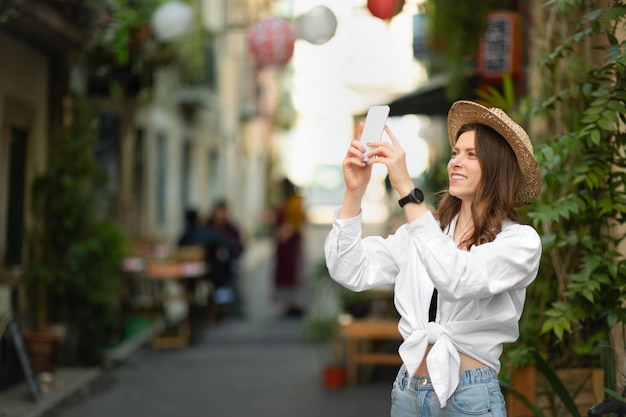 Geïnspireerde gelukkige jonge blanke dame in casual en hoed maakt foto op smartphone kijkt naar bezienswaardigheden in nieuwe stad