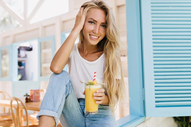 Geïnspireerde vrouw met lang kapsel met glas sap en poseren met een glimlach Portret van gebruinde blonde model in spijkerbroek zittend op de vensterbank en cocktail drinken