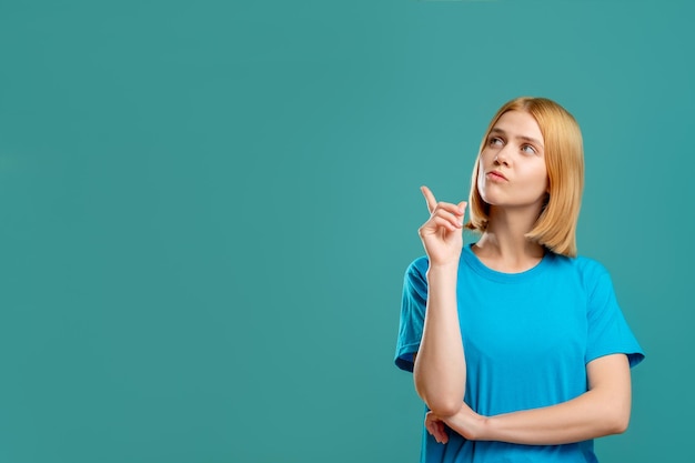 Geïnspireerde vrouw portret antwoord oplossing Peinzende blonde dame in blauwe t-shirt wijzend op lege ruimte voor logo tekst geïsoleerd op groenblauw promotionele achtergrond Insight idee