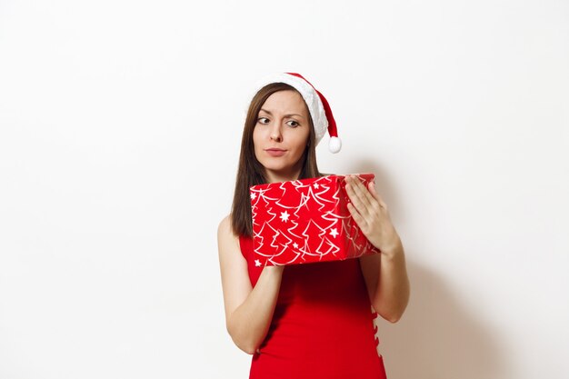 Geïnteresseerde en verloofde jonge vrouw gekleed in rode jurk en kerstmuts houdt geschenkdoos, denkt dat ze op een witte achtergrond is gegeven. Santa meisje met heden geïsoleerd. Nieuwjaarsvakantie 2018 concept