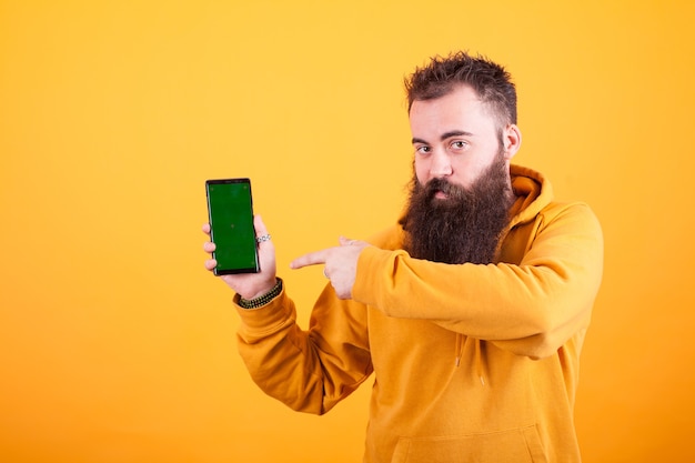 Geïntrigeerde bebaarde hipster man wijzend op smartphone over gele achtergrond. Gele hoodie. Knappe man. Moderne technologie