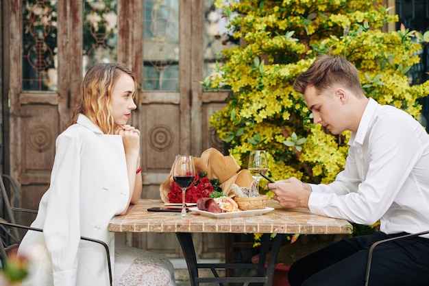 Geïrriteerde jonge vrouw die vriendje bekijkt dat vrienden sms't of sociale media controleert in plaats van met haar te praten tijdens een romantische date