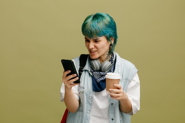 Geïrriteerde jonge vrouwelijke student met koptelefoon en bandana op nek en rugzak met papieren koffiekopje met behulp van mobiele telefoon geïsoleerd op olijfgroene achtergrond