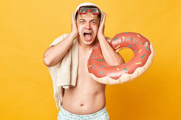 Geïrriteerde volwassen man met korte zwembroek zwembroek snorkelbril met donut rubberen ring en zak geïsoleerd op gele achtergrond schreeuwend depressief zijn oren bedekken met mond