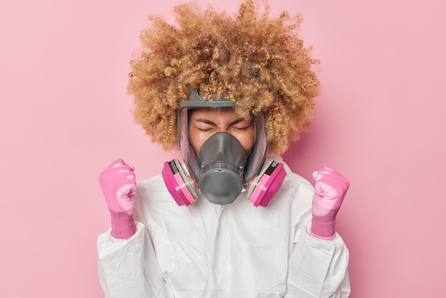 Geïrriteerde vrouw met krullend borstelig haar draagt een gasmasker gekleed in wit beschermend pak en handschoenen balt vuisten verkent omgeving maatregelen vervuiling geïsoleerd over roze achtergrond