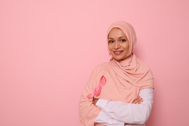 Geïsoleerd portret van arabische moslim mooie vrouw die roze hijab draagt met roze satijnen lint in symbool van de internationale dag van kankerbewustzijn. oncologische ziekten concept. ruimte kopiëren op roze achtergrond