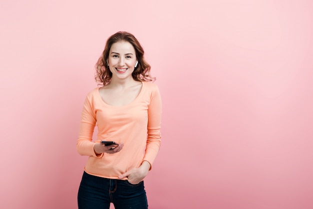 Geïsoleerd portret van glimlachende jonge vrouw die mobiele telefoon in hand houden, draadloze hoofdtelefoons dragen, die toevallig overhemd dragen