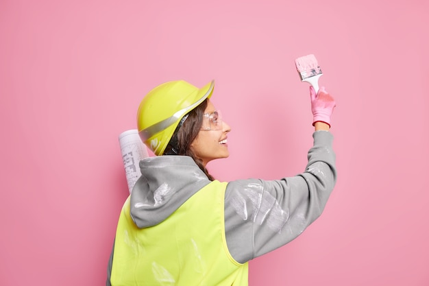 Geïsoleerd schot van vrolijke bekwame vrouwelijke bouwvakker staat terug verven muur in roze kleur maakt gebruik van kwast houdt bouwblauwdruk draagt helm en uniform. Onderhoudsdienst