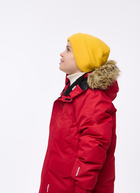 Geïsoleerd zijportret van een knap kind, schattige preteen schooljongen in felgele wollen muts en rood donsjack met besneeuwde capuchon voor hem kijkend op kopie ruimte van witte achtergrond