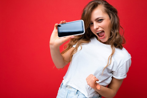 Gek lachende jonge blonde vrouw goed uitziende dragen van wit t-shirt staande geïsoleerd op rode achtergrond met kopie ruimte met telefoon weergegeven: smartphone in de hand met leeg scherm voor mockup lo