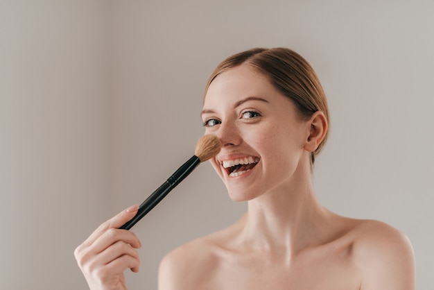 Gek worden. Aantrekkelijke jonge vrouw die naar de camera kijkt en haar neus aanraakt met een make-upborstel terwijl ze tegen de achtergrond staat