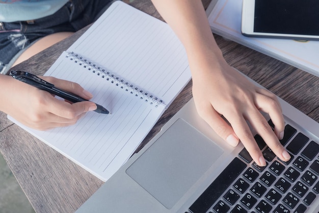 Gekapte afbeelding van een vrouw die op een boek schrijft terwijl ze een laptop aan tafel gebruikt