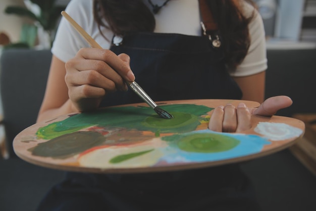 Gekapte afbeelding van een vrouwelijke kunstenaar die voor een easel staat en een penseel in een kleurenpalet dompelt