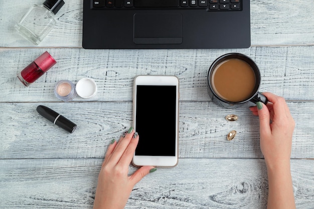 Gekapte hand van een vrouw die koffie vasthoudt terwijl ze een telefoon op tafel gebruikt