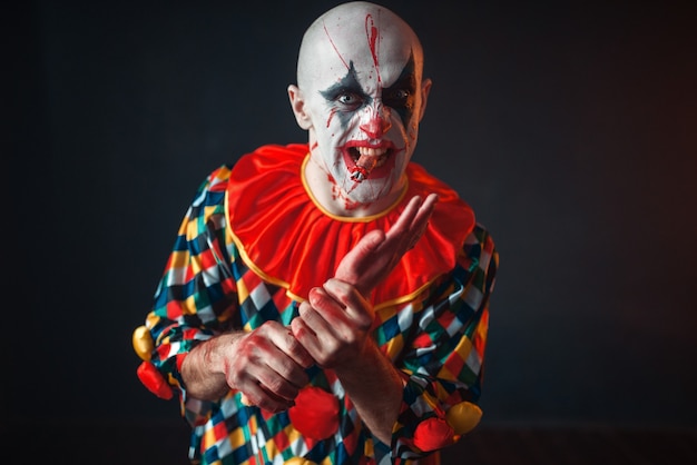 Gekke bloedige clown houdt menselijke hand, vinger in zijn tanden. Man met make-up in halloween-kostuum, gekke maniak
