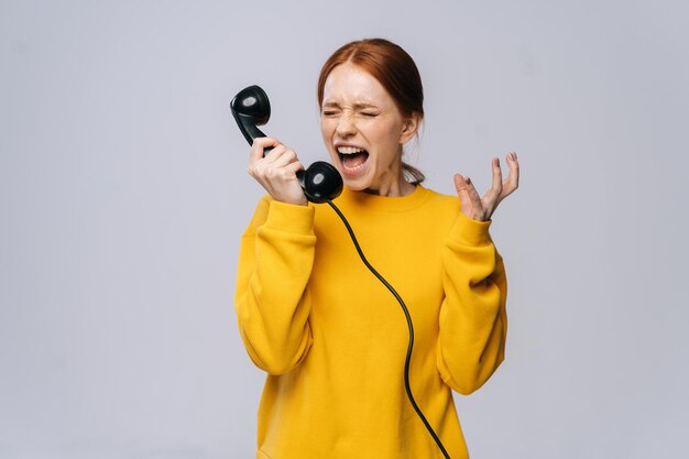 Gekke boze jonge vrouw in stijlvolle gele trui praten over retro telefoon en schreeuwen in handset