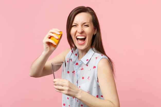 Gekke lachende jonge vrouw in zomerkleren houdt de helft van vers rijp oranje fruit vast, glazen beker geïsoleerd op roze pastel muur achtergrond. Mensen levendige levensstijl ontspannen vakantie concept. Bespotten kopie ruimte.