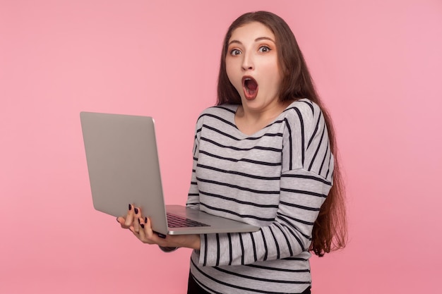 Gekke online studiecursus Portret van verbaasd verraste vrouw in gestreept sweatshirt die laptop vasthoudt en kijkt met geschokte uitdrukking die op computer studio opname werkt geïsoleerd op roze achtergrond