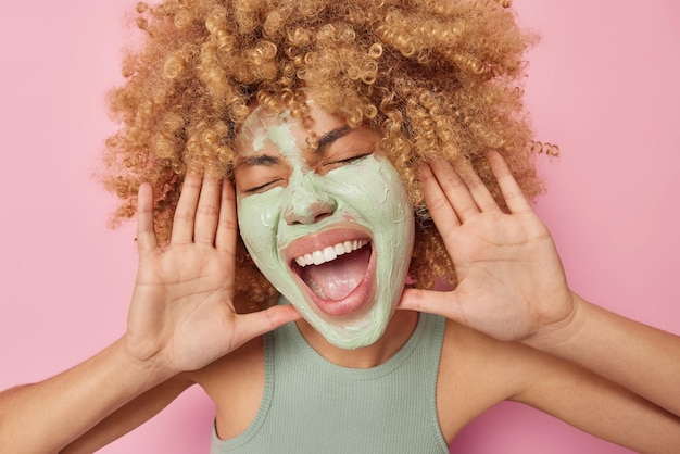 Gekke vrouw met krullend haar houdt handpalmen in de buurt van gezicht roept luid houdt mond wijd geopend gesloten ogen past schoonheid gezichtsmasker toe draagt casual t-shirt geïsoleerd over roze achtergrond Huidverzorging