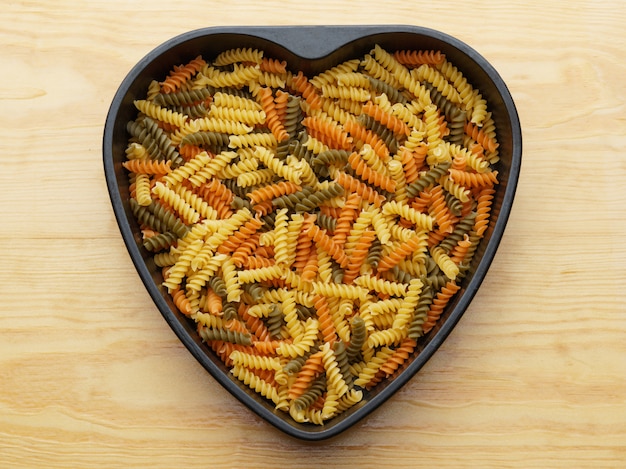 Gekleurde pasta in een metalen schotel in de vorm van een hart, bovenaanzicht.